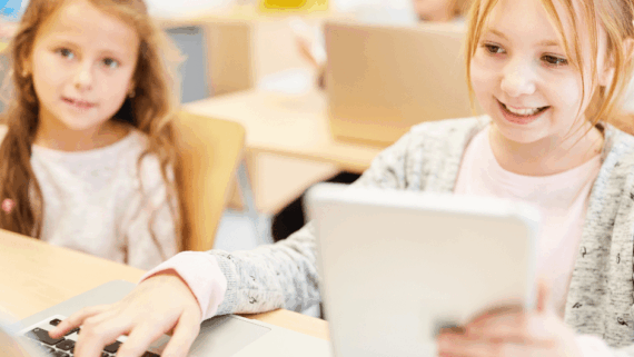 Dues nenes en primer pla en una aula. Una està treballant amb una llibreta i l'ordinador. En aquest context cal cuidart la salut ocular