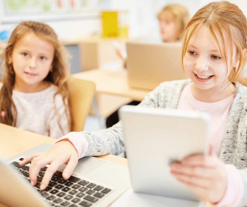 Dues nenes en primer pla en una aula. Una està treballant amb una llibreta i l'ordinador. En aquest context cal cuidart la salut ocular